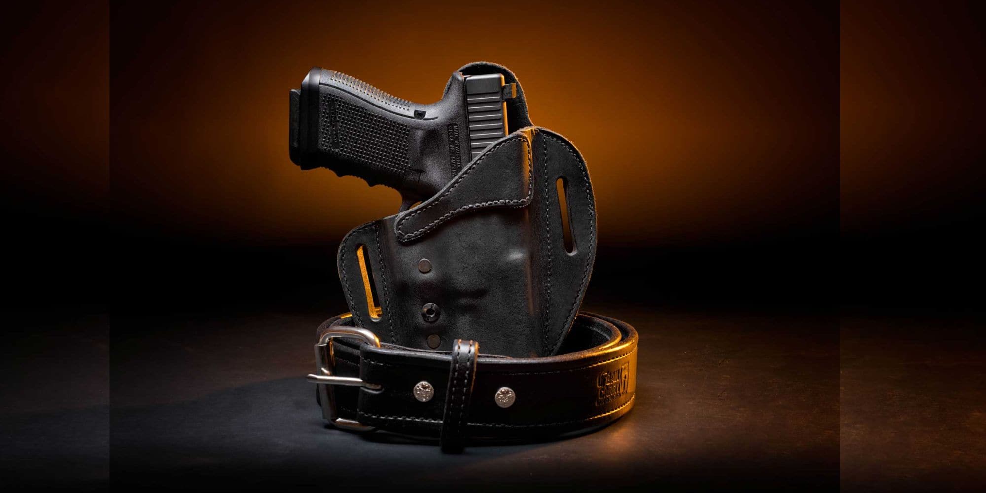 Black leather outside-the-waistband holster with a handgun, displayed with a sturdy Urban Carry gun belt on a dark background with warm lighting.