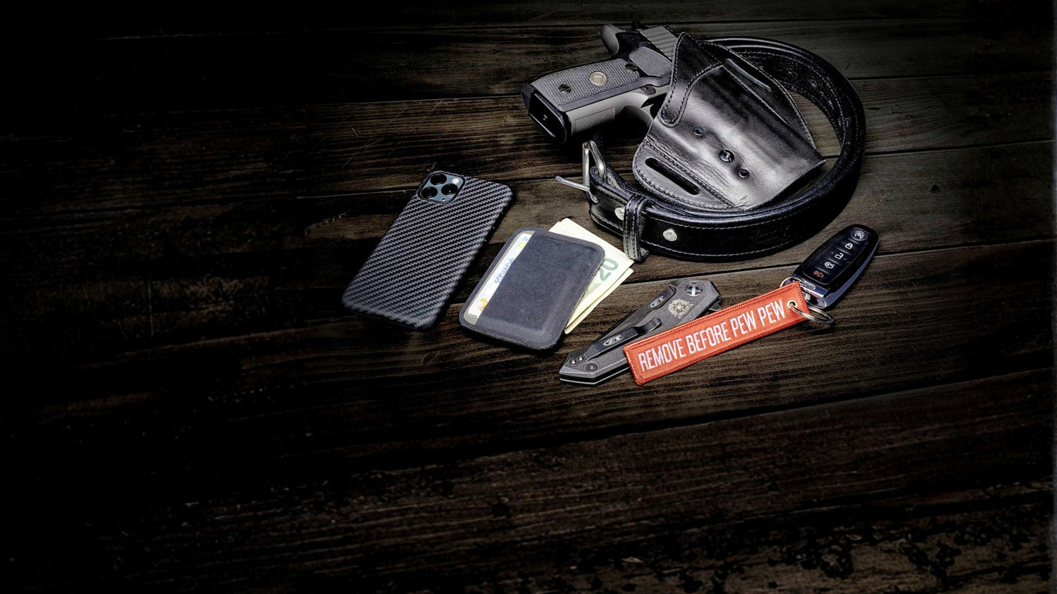 Collection of items including a gun, holster, phone, wallet, and knife on a dark wooden surface.