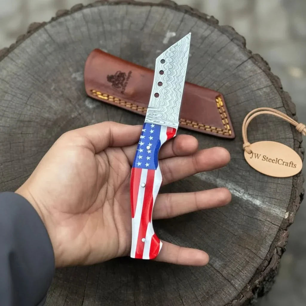 Patriotic fixed blade knife with Damascus pattern held in hand. Leather sheath in background on a tree stump, branded 'JW SteelCrafts'.