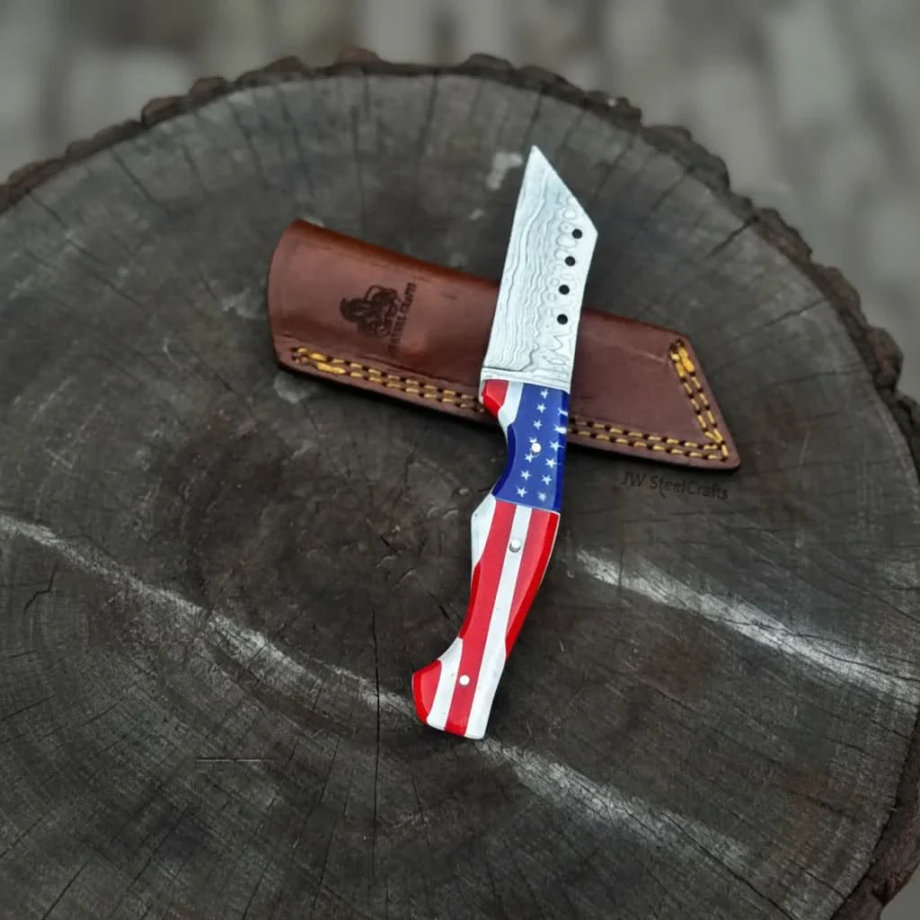 Patriotic fixed blade knife with Damascus pattern sitting on a leather sheath on a tree stump, branded 'JW SteelCrafts'.