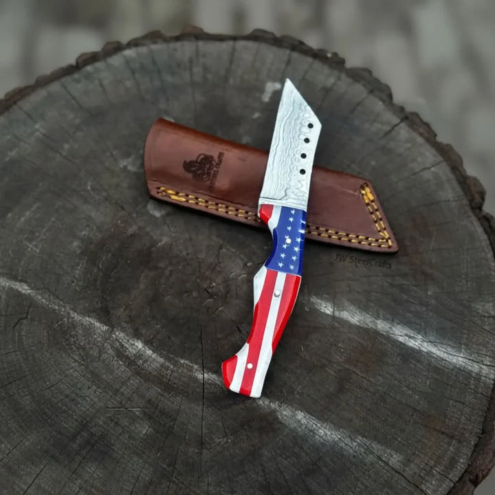 Patriotic fixed blade knife with Damascus pattern sitting on a leather sheath on a tree stump, branded 'JW SteelCrafts'.