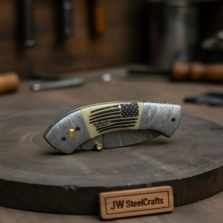 Folding knife with American flag design on a wooden surface, branded 'JW SteelCrafts'.