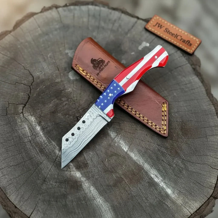 USA Patriotic fixed blade knife with Damascus pattern sitting on a leather sheath on a tree stump, branded 'JW SteelCrafts'.